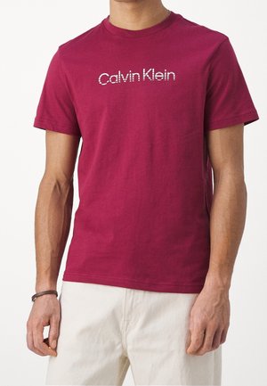Mężczyzna ubrany w bordowy t-shirt Calvin Klein z krótkim rękawem oraz jasne spodnie, z ciemną opaską na lewej ręce.