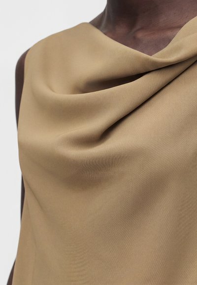 Κοντινό σε άτομο που φοράει μπεζ αμάνικη μπλούζα με ντραπαριστό ντεκολτέ, δείχνοντας την υφή του υφάσματος και την αντίθεση του τόνου του δέρματος.