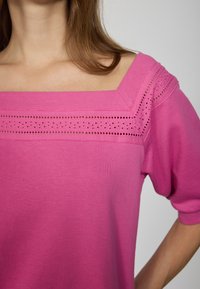 Žena v růžovém topu s čtvercovým výstřihem, zdobeném krajkovým lemováním kolem výstřihu a balónkovými krátkými rukávy.