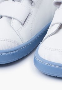 Weiße Sneakers mit strukturiertem Obermaterial und blauer Gummisohle, ausgestattet mit Klettverschlüssen, die ein minimalistisches Design und kontrastierende Farben betonen.