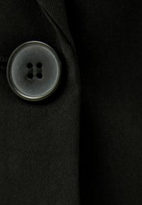 Svart tyg med en närbild av en cirkulär knapp, med fyra hål och en slät yta, mot en texturerad bakgrund.