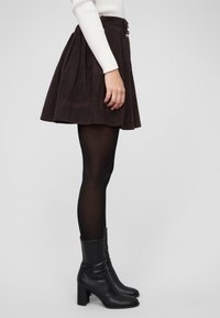 Mujer con una falda marrón de pana, medias negras transparentes, botines negros de tacón y una camiseta blanca de manga larga con canalé, de perfil.