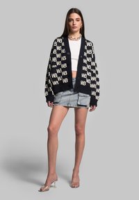 Námořnický pletený cardigan s opakujícím se off-white vzorem "WB", ve spojení s bílým crop topem a denimovou mini sukní. Průhledné muly na podpatku.