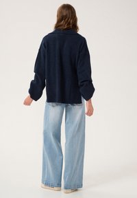 Pulover bleumarin cu guler, cu o croială lejeră și tiv cu nervuri, purtat cu blugi deschis la culoare, cu croială largă, arătând o vedere din spate casual.