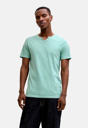 Homme portant un t-shirt henley à manches courtes vert clair et un pantalon large foncé, debout devant un fond blanc.