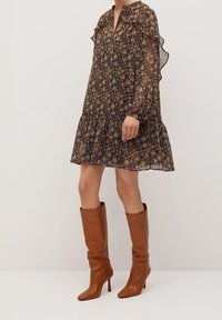 Femme portant une longue robe florale marron à manches longues avec des détails en volants, associée à des bottes en cuir beiges à talons montant jusqu'aux genoux.