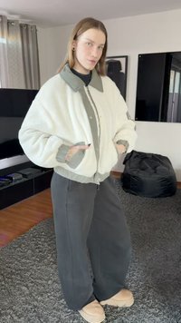 Witte, gestructureerde bomberjack met een grijze kraag en manchetten, gecombineerd met losse grijze broek en beige schoeisel. Eenvoudig, functioneel ontwerp.