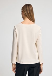 Langärmliges Oberteil in hellbeige, aus weicher Baumwolle. Hat einen runden Ausschnitt und eine lockere Passform mit gestepptem Saum.