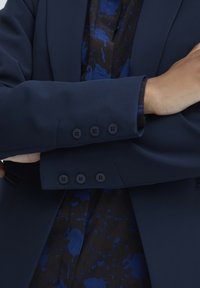 Blazer bleu marine au design structuré, avec une texture lisse et quatre boutons noirs sur les poignets. En dessous, une chemise à motifs bleu foncé.