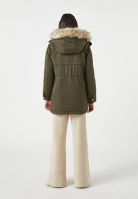 Olijfgroene parka met een met bont afgewerkte capuchon, voorzien van een taillekoord en een sluiting met knopen aan de voorkant. Draag het met lichtbeige wijde broekspijpen.