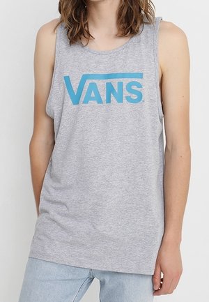 Persoon die een grijs mouwloos tanktop draagt met een blauw "VANS"-logo en lichtblauwe spijkerbroek, staand tegen een effen witte achtergrond.