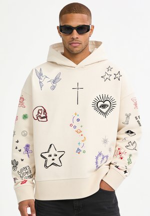 Man wearing beige hoodie with colorful doodles including stars, birds, skull, heart with eye, and other symbols, paired with black sunglasses.