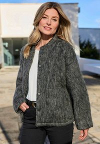 Grijze, gestructureerde sweaterjacket met een ronde kraag, drukknoopjes en voorzakken. Gecombineerd met een witte top en zwarte broek.
