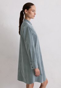 Vrouw met bruin haar in een paardenstaart, gekleed in een loszittende, langmouwige, lichtblauwe blousejurk tegen een witte, gestructureerde muur.