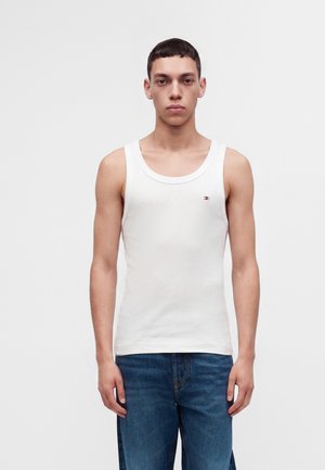 Jeune homme portant un débardeur blanc sans manches et un jean bleu, debout devant un fond blanc uni.