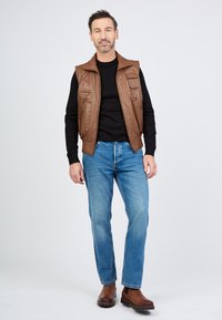Brauner Lederweste mit mehreren Taschen über einem schwarzen Hemd, kombiniert mit hellblauen Jeans und braunen Stiefeln. Neutrale Hintergrund.
