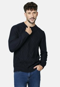 Suéter de punto azul marino con patrones de diamante, puños y dobladillo acanalados. Cuello redondo con detalles texturizados. Corte casual, usado sobre unos jeans azules.