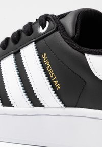 Mustat nahkakengät, joissa on kolme valkoista raitaa, kultaiset "SUPERSTAR"-tekstit, pehmustettu nilkkakuppi ja valkoinen kumipohja.