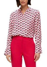 Långärmad skjorta med ett rosa och svart pilmönster på en vit bakgrund. Har en krage och utsvängda manschetter. Matchas med röda byxor.