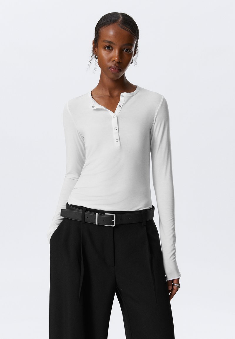 Witte langemouwen henley-top met knoopsluiting, gecombineerd met hoge zwarte broek en een zwarte riem. Gladde stoftextuur.