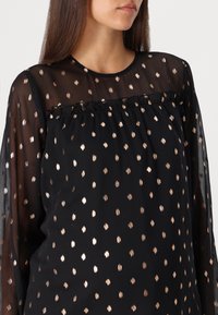 Blouse noire avec manches longues transparentes, ornée de motifs à pois dorés et d'un col à volants. Tissu léger avec une finition texturée.
