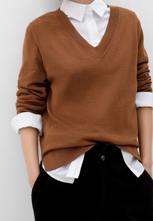 Person iført en brun V-hals sweater over en hvid skjorte med opkrogede ærmer og sorte bukser, hænderne i lommerne.