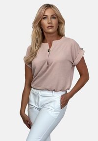 Bluzka z krótkim rękawem w odcieniu blush różu z okrągłym dekoltem i guzikami na podwiniętych mankietach, w zestawieniu z dopasowanymi białymi spodniami. Gładka tekstura.