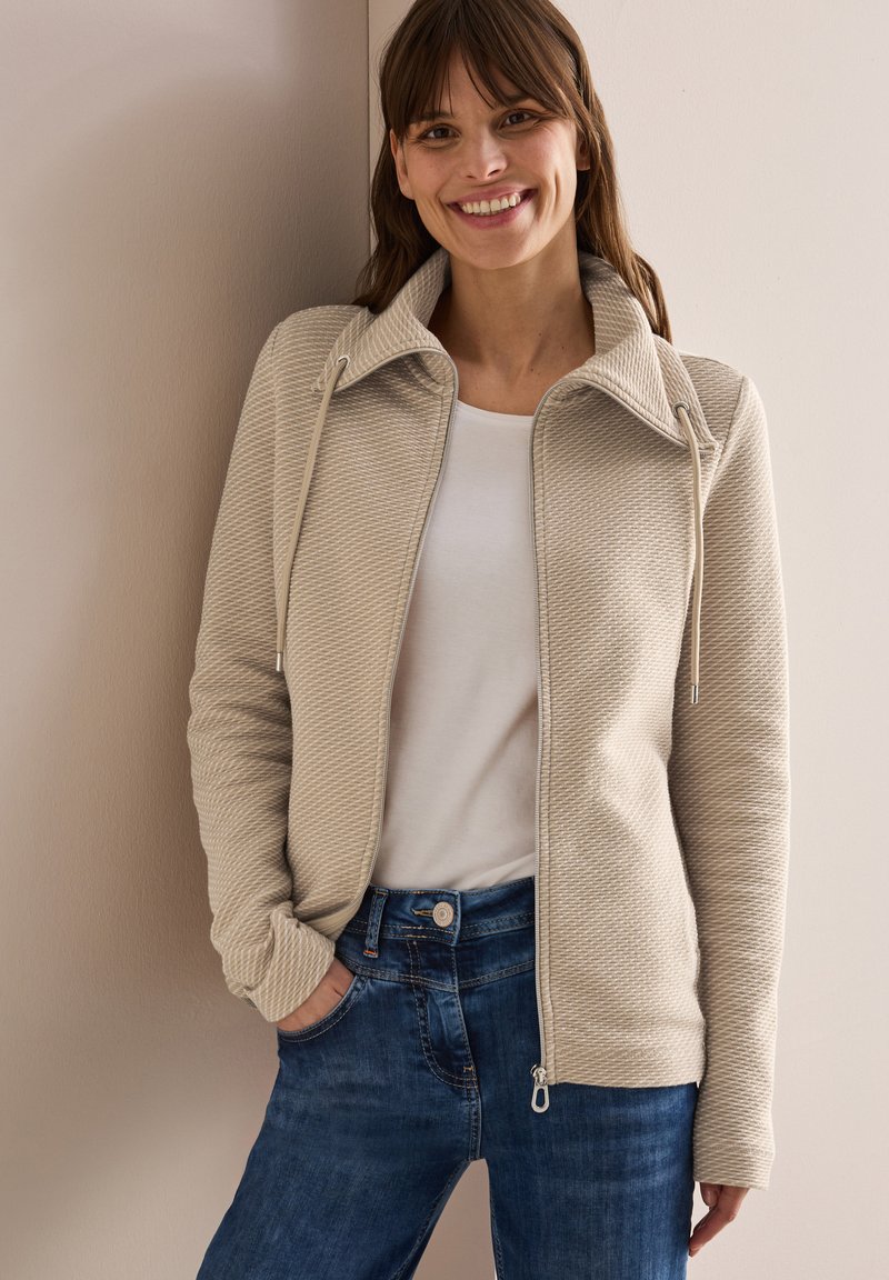 Chaqueta beige con cremallera y acabado texturizado, con cuello y cordones, combinada con una camiseta blanca y jeans de mezclilla azul.