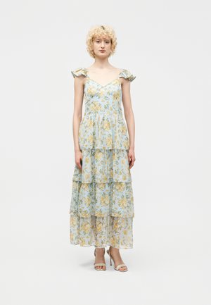 Γυναίκα με κατσαρά ξανθά μαλλιά που φοράει μπλε μακρύ φόρεμα με πολλαπλές στρώσεις και φλοράλ μοτίβα, με βολάν στους ιμάντες στους ώμους και μπεζ ψηλοτάκουνα σανδάλια, όρθια.