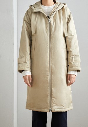 Femme portant un long manteau matelassé beige avec une capuche, une fermeture éclair frontale et des détails de sangle sur un pull en maille blanc et un pantalon noir.