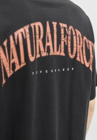Black cotton t-shirt featuring a large, cracked orange graphic text "NATURALFORCE" and smaller white text "NEVERSLEEP" below it.