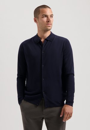 Navy long-sleeve button-up shirt in textured fabric, with a pointed collar and dark buttons, paired with neutral trousers.