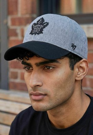 Junger Mann mit kurzen lockigen Haaren, der eine graue und schwarze Toronto Maple Leafs Baseballmütze trägt, vor einer Ziegelwand sitzend.