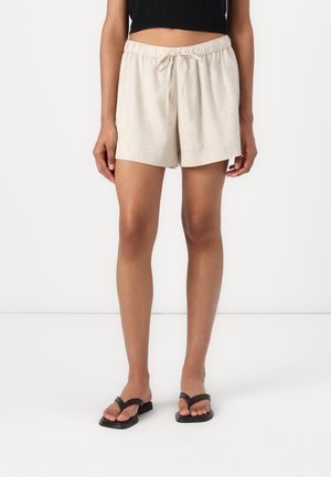 Kvinde iført beige linnedragt med snøre i taljen, sort ærmeløs crop-top og sorte flade sandaler, stående mod en hvid baggrund.