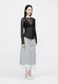 Čierne priehľadné tričko s dlhým rukávom s textúrovanými bodkami, v kombinácii s svetlosivou sukňou s čipkovaným lemom a bielymi mule obuvami so špicatou špičkou.