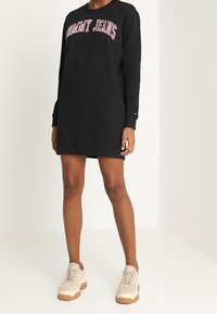 Musta collegepaidan mekko, jossa on punainen "TOMMY JEANS" -logo rinnassa. Mekossa on pitkät hihat ja rento istuvuus. Yhdistetty vaaleanbeigeihin lenkkareihin.