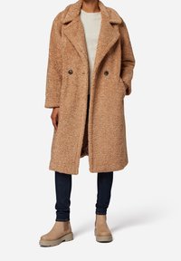 Lichtbruine, luxueuze, dubbelgeknoopte jas met grote revers, zijkanten zakken en twee zwarte knopen; gecombineerd met donkerblauwe spijkerbroek en beige enkellaarzen.