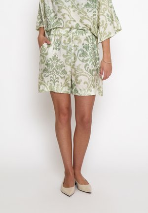 Model iført hvide shorts med grønt blomsterprint og matchende top, parret med beige spidse sko, mod en ensfarvet baggrund.
