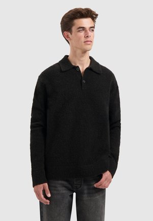 Jongeman met kort bruin haar, gekleed in een zwarte lange mouwen pullovertrui met kraag en twee knopen, gecombineerd met donkergrijze jeans.