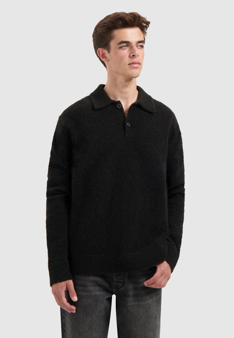 Jongeman met kort bruin haar, gekleed in een zwarte lange mouwen pullovertrui met kraag en twee knopen, gecombineerd met donkergrijze jeans.