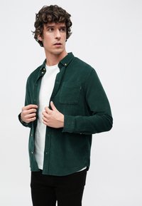 Camicia verde di velluto a coste con colletto abbottonato, taschino sul petto e cuciture visibili. Indossata sopra una t-shirt bianca, con pantaloni neri.