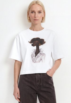 Mujer rubia vestida con camiseta corta blanca con ilustración negra de caniche en blusa con volantes, combinada con vaqueros oscuros de talle alto.