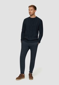 Marineblauwe kabelgebreide trui met geribbelde manchetten en onderrand, gecombineerd met marineblauwe taperende broek en bruine veterschoenen met textuurelementen.