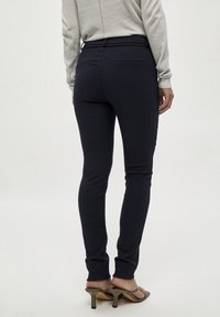 Pantaloni attillati blu navy con una texture liscia, dotati di una cintura e tasche posteriori, abbinati a una maglietta grigia chiara a maniche lunghe e tacchi.