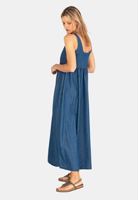 Maxi jurk in denim blauw met een getailleerde bodice en een vloeiende rok. Kenmerkt brede schouderbanden en een gathering in de taille, gecombineerd met platte sandalen.
