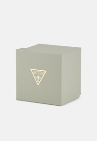 Green textured cardboard box with a triangular gold logo on the front, featuring the word "GUESS" and a question mark design.