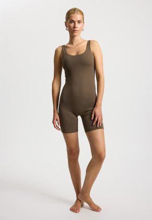 Žena stojící naboso, oblečená v přiléhavém bezrukávovém zelenohnědém overalovém kostýmku s kraťasy na jednobarevném světle pozadí.