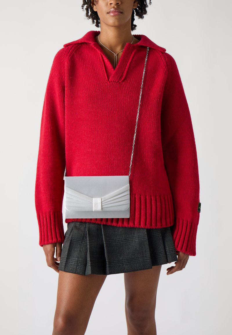 Maglione rosso lavorato a maglia con scollo a V e polsini a coste, abbinato a una clutch grigia con parte anteriore plissettata e tracolla a catena, su una gonna plissettata.