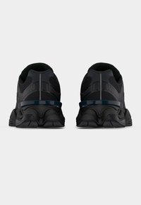 Ζευγάρι μαύρα αθλητικά παπούτσια εμφανιζόμενα από πίσω με παχιά σόλα και ανακλαστικές λωρίδες στην περιοχή της φτέρνας, πάνω σε απλό φόντο.