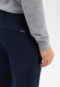 Pantalon marine en tissu doux, avec une poche arrière ornée d'un discret détail métallique. La main du modèle repose sur la jambe, portant un pull gris.